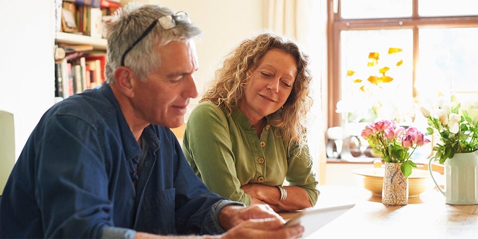 Middle-aged couple sitting at kitchen counter reviewing documents on a laptop.