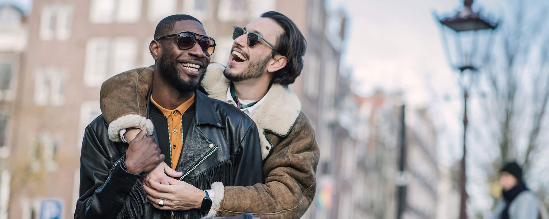 Same-sex couple embraced on street in late autumn or early winter in Amsterdam. Caucasian and African ethnicity, brown hair, sunglasses, wearing leather jackets.