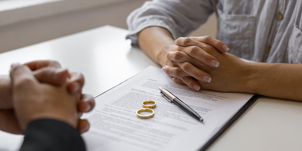 Two people sitting at a table, hands folded, with paperwork, a pen and wedding rings between them.