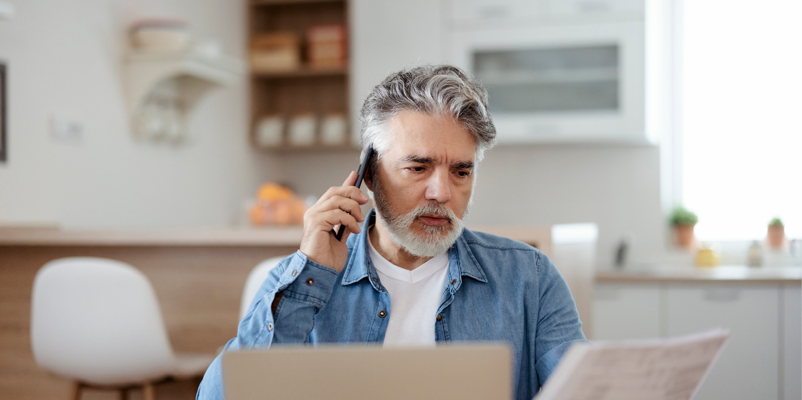 Man talking on phone while looking at papers in front of a laptop