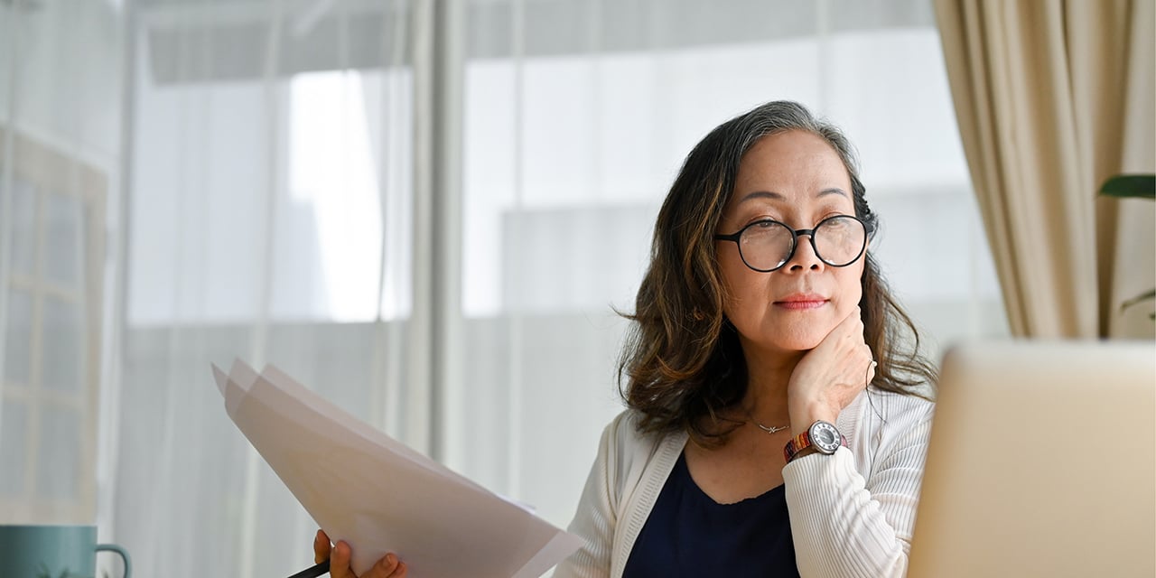 Woman wearing glasses while looking at a computer and holding papers.