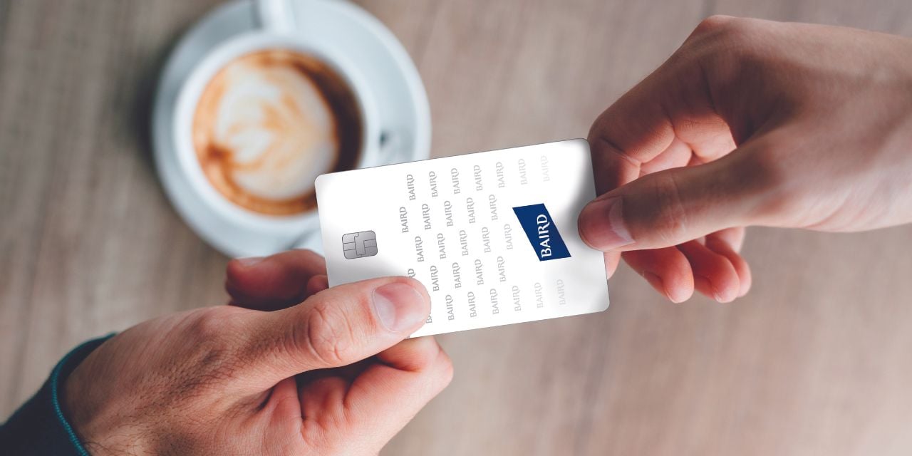 Two hands exchanging a microchipped card printed with the Baird logo over a cup of coffee on a table