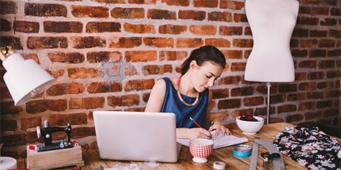 Brunette woman, sitting and sketching. She is surround by fabrics, thread, a dress form and other sewing materials.