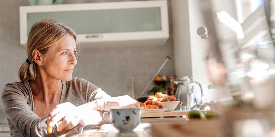 Older, blonde woman sitting at table with mug of coffee looking out the window.